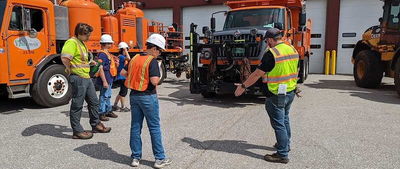 Youth and AOT Staff with hard hats and reflective vests in from of equipment and trucks