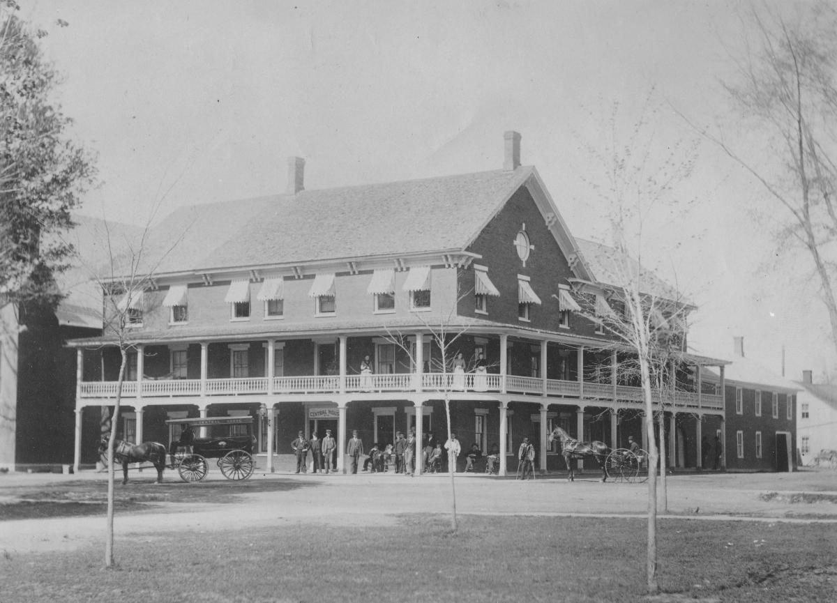 A black and white image of the Eagle House taken in 1890. Image feature women and men dressed in clothes of the era standing at the entrance of the building as horse-drawn carriages amble by.