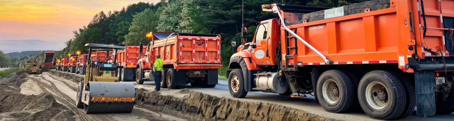 Large orange VTrans dump trucks sit in a long line next to a dug up roadway.